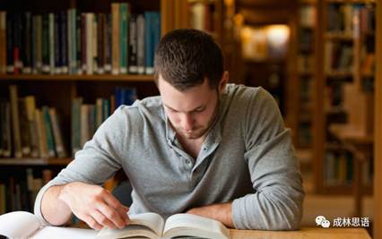 A young man is reading a book at a table in a library surrounded by shelves filled with books.